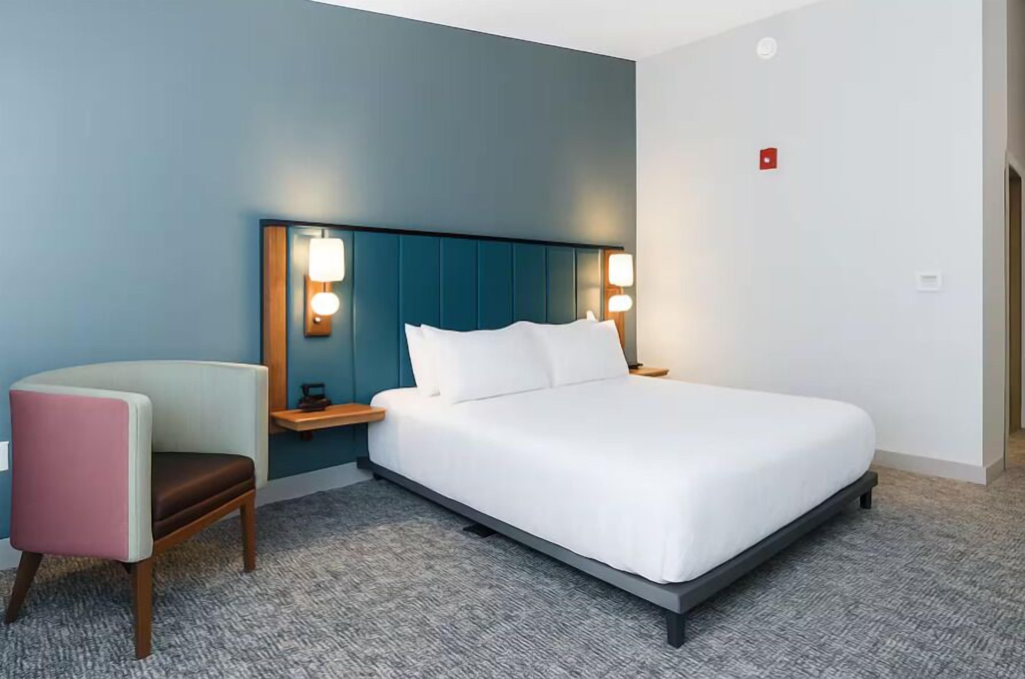 Una habitación de hotel moderna con cama, mesitas de noche, una silla y cabecero con iluminación integrada.