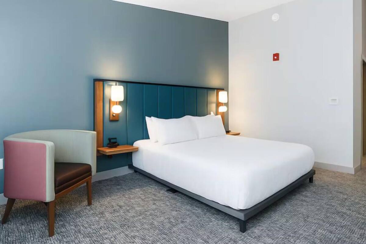 Ein modernes Hotelzimmer mit Bett, Nachttischen, einem Sessel und einem Kopfteil mit integrierter Beleuchtung.