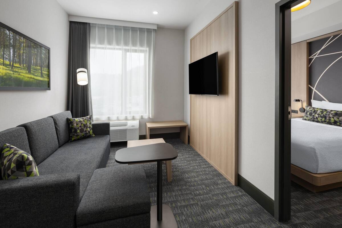 Ein modernes Hotelzimmer mit einer Couch, einem kleinen Tisch, einem Fernseher, einem Bett und einem Fenster mit Vorhängen.