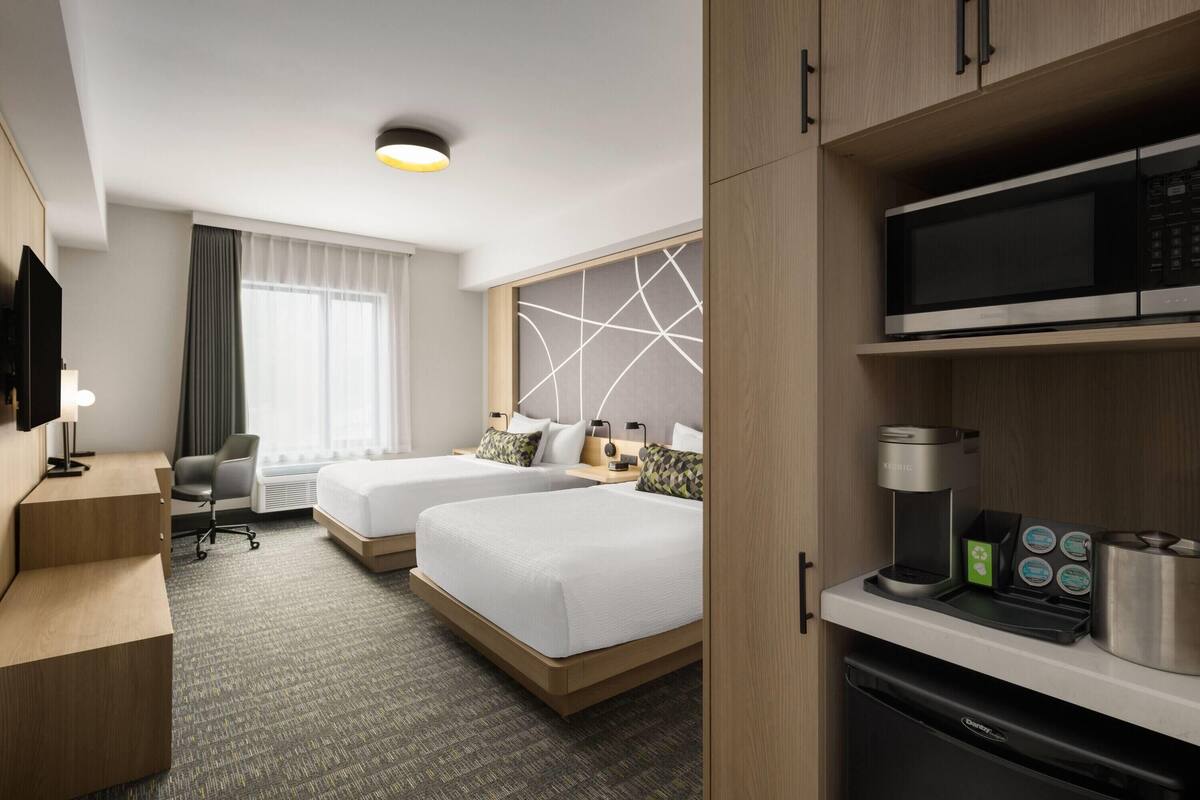 Ein Hotelzimmer mit zwei Betten, einem Schreibtisch, einem Stuhl, einem Fernseher, einer Kaffeemaschine und einer Mikrowelle.