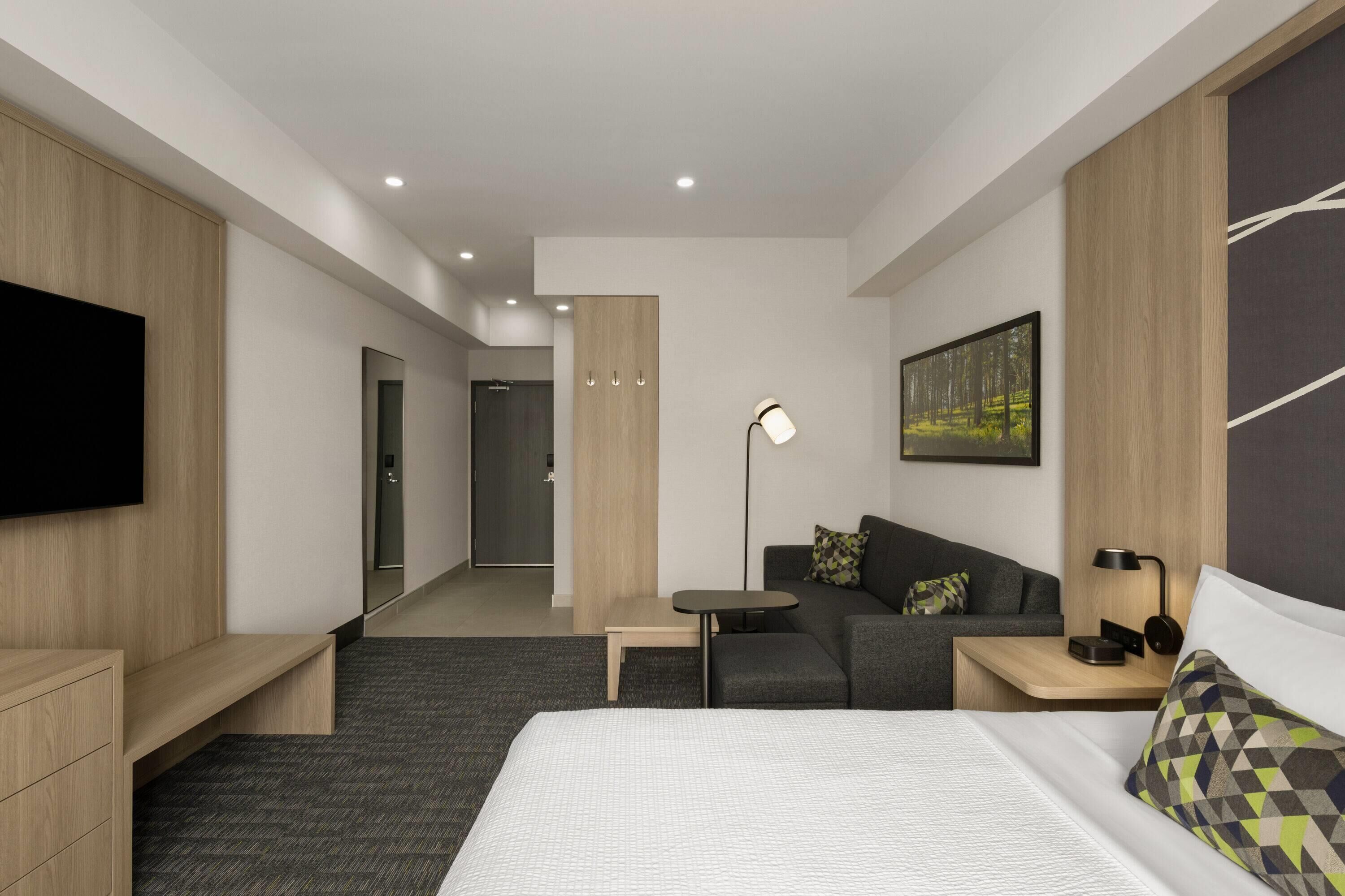 A modern hotel room with a bed, a sofa, a desk, a flat-screen TV, a floor lamp, and a painting on the wall.