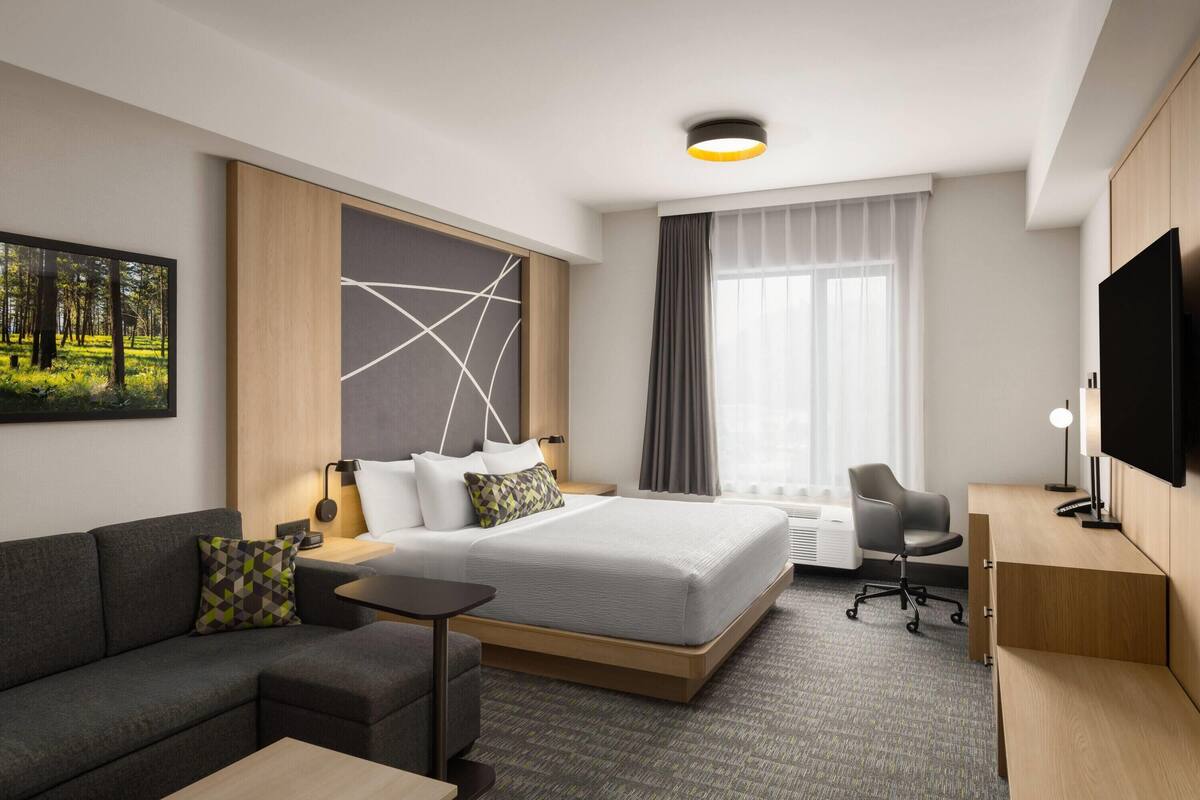 Ein modernes Hotelzimmer mit einem Bett, einer Couch, einem Schreibtisch, einem Fernseher und einem großen Fenster.