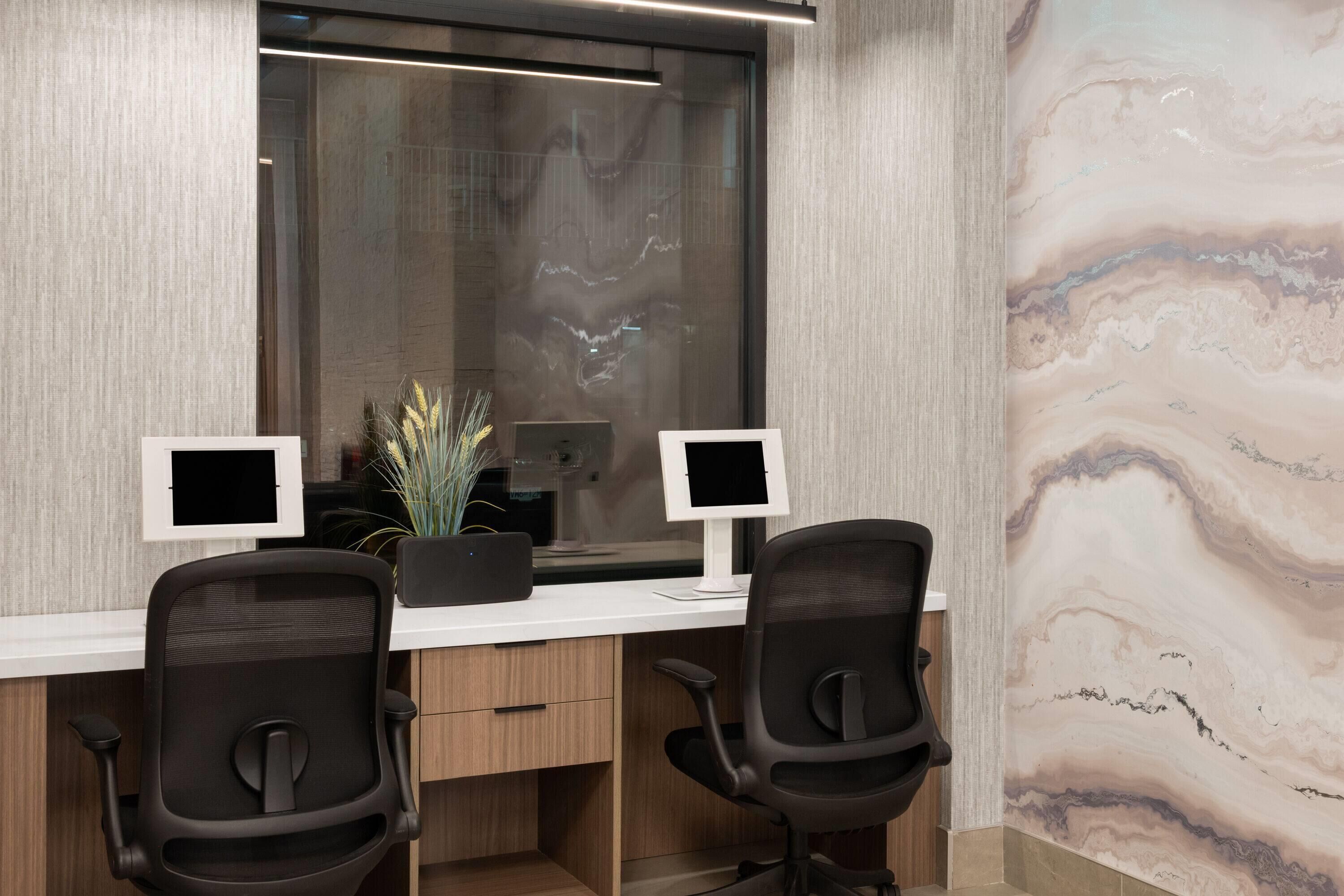 Un espacio de oficina moderno con dos escritorios, cada uno con un monitor de computadora, una planta en maceta y una silla.