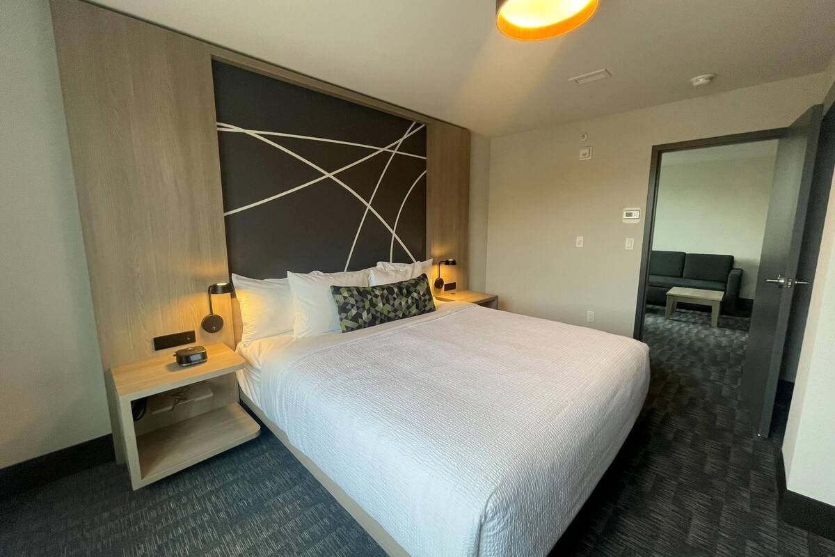 Ein modernes Hotelzimmer mit einem großen Bett, Nachttischen, einer Wandleuchte und einer Tür zu einem weiteren Raum.