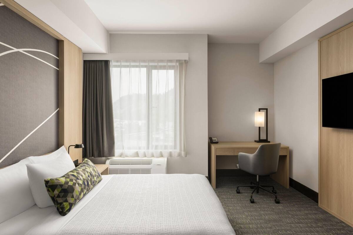 Ein Hotelzimmer mit Bett, Schreibtisch, Stuhl, Fernseher und einem Fenster mit Vorhängen.