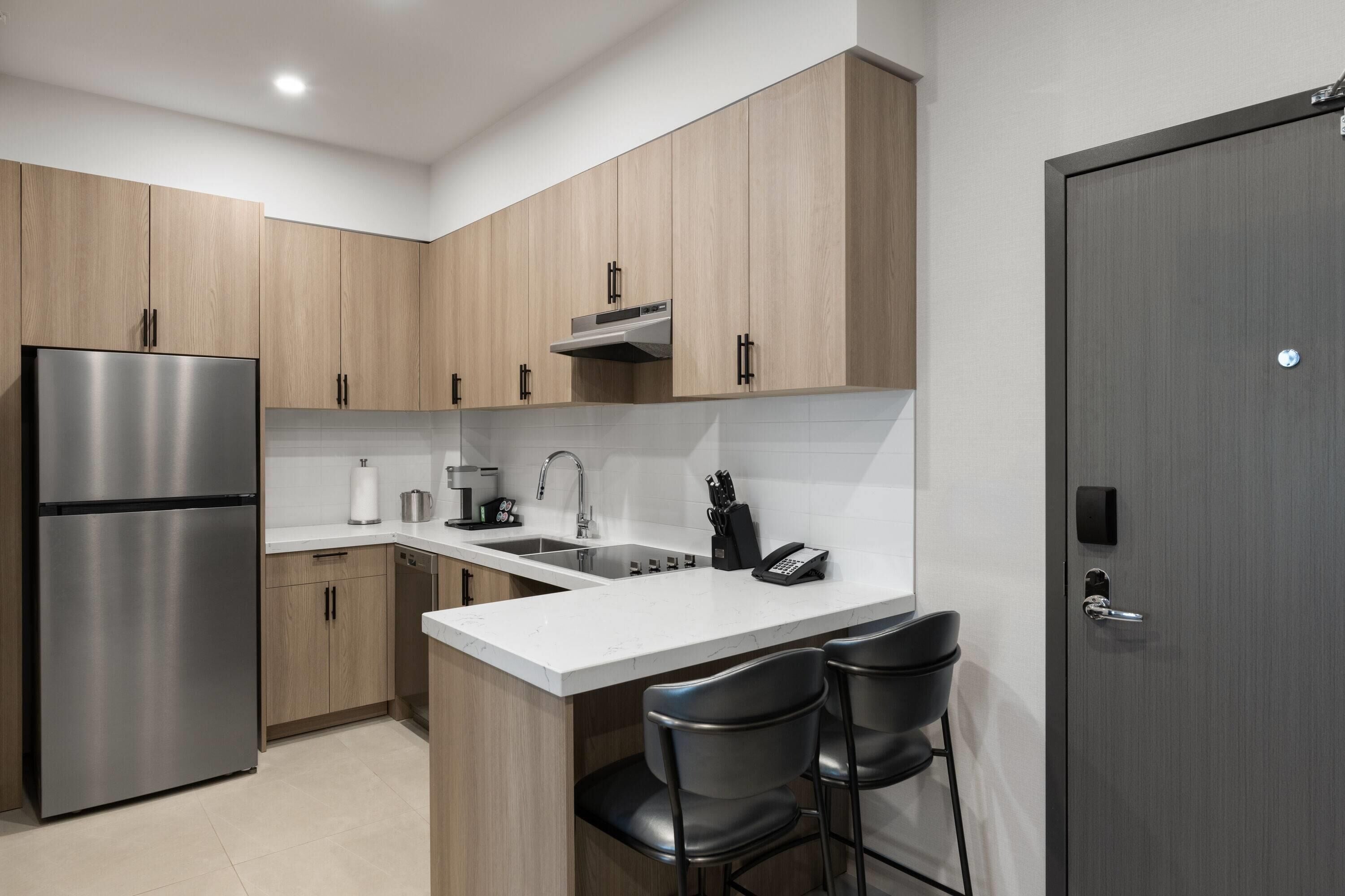 Una cocina moderna con un refrigerador de acero inoxidable, armarios de madera, una encimera blanca, un lavamanos y dos taburetes de bar.