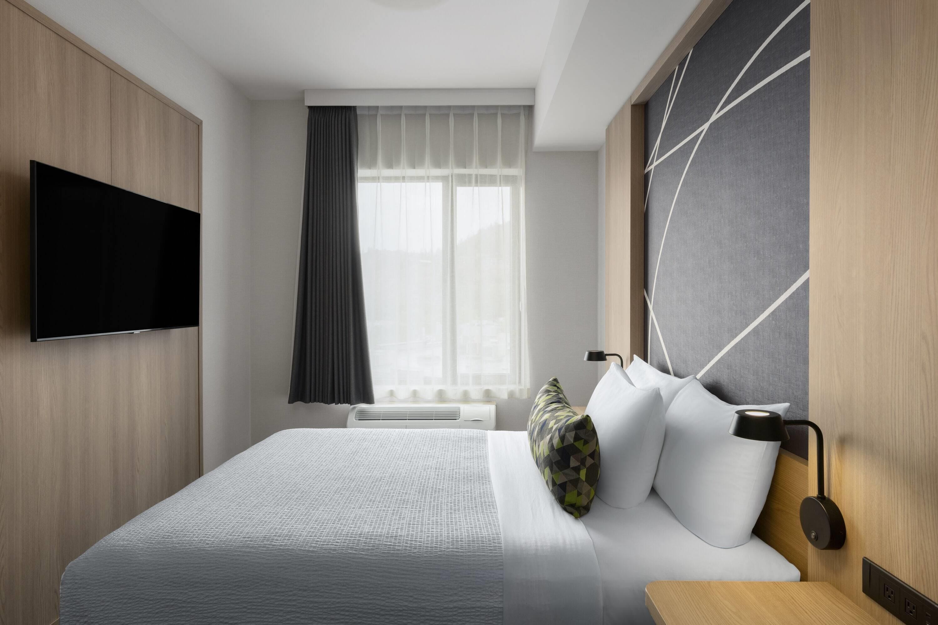 A modern hotel room with a flat-screen TV, a large bed with white bedding, a wall-mounted lamp, and a window with sheer curtains.