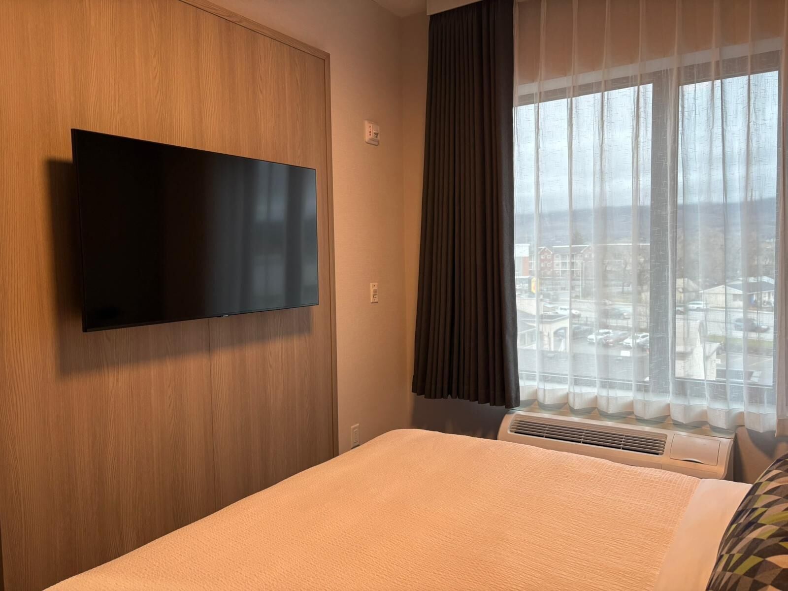 A hotel room with a bed, a flat-screen TV mounted on a wooden wall, a window with sheer curtains, and an air conditioning unit.