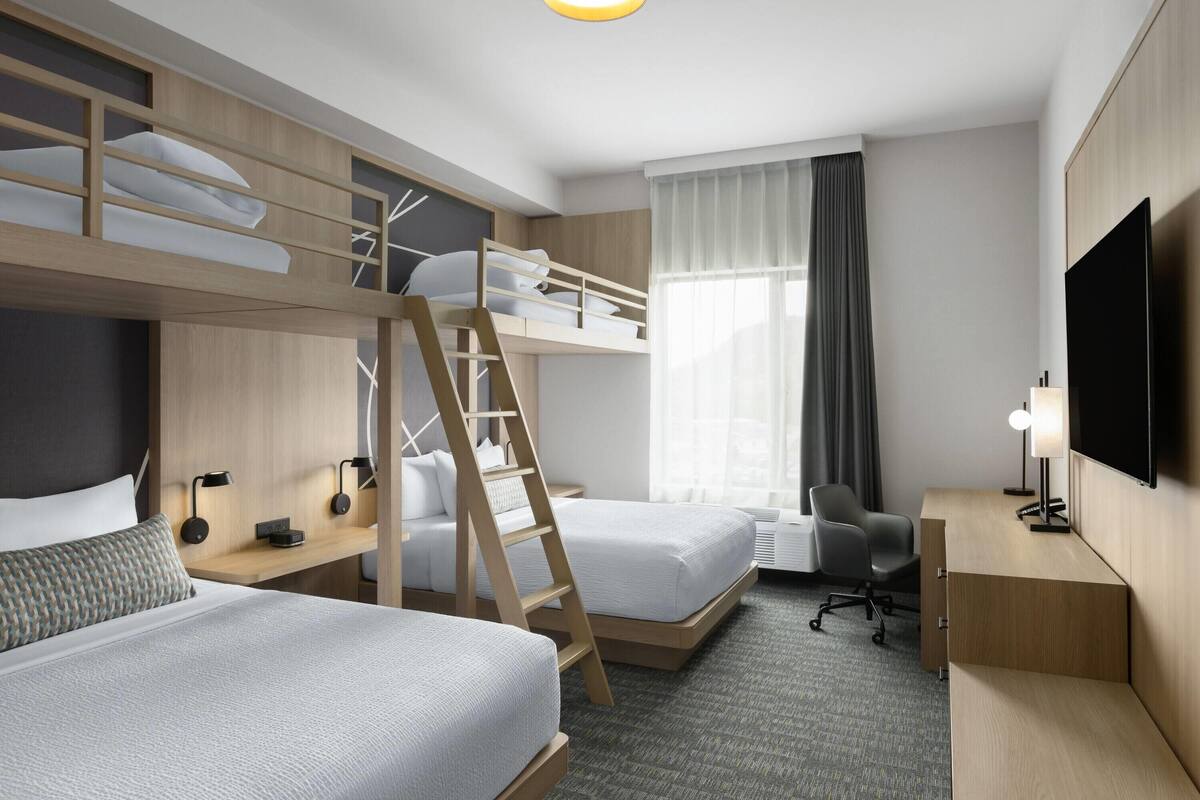 Ein Hotelzimmer mit Etagenbett, Schreibtisch, Stuhl, Fernseher und einem Fenster mit Vorhängen.