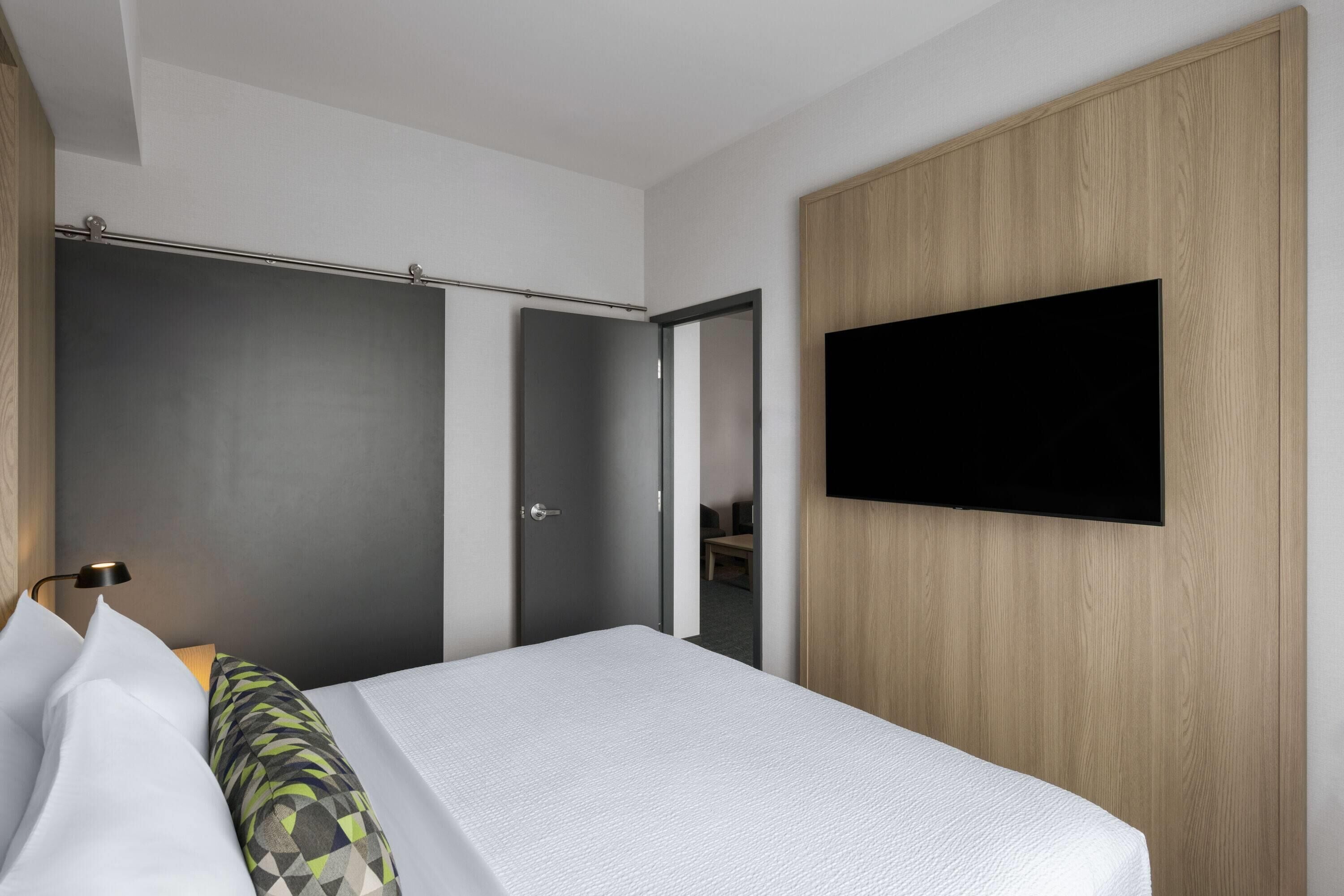 A modern hotel room with a bed, a flat-screen TV mounted on the wall, a wooden wardrobe, and a door leading to another room.