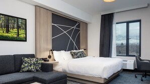 A modern hotel room with a large bed, a sofa, a desk, and a view of a snowy landscape through a large window. - La Quinta Inn & Suites By Wyndham Kelowna Bc (Kelowna)