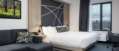 Une chambre d’hôtel moderne dotée d’un grand lit, d’un canapé, d’un bureau et offrant une vue sur un paysage enneigé par une grande fenêtre.