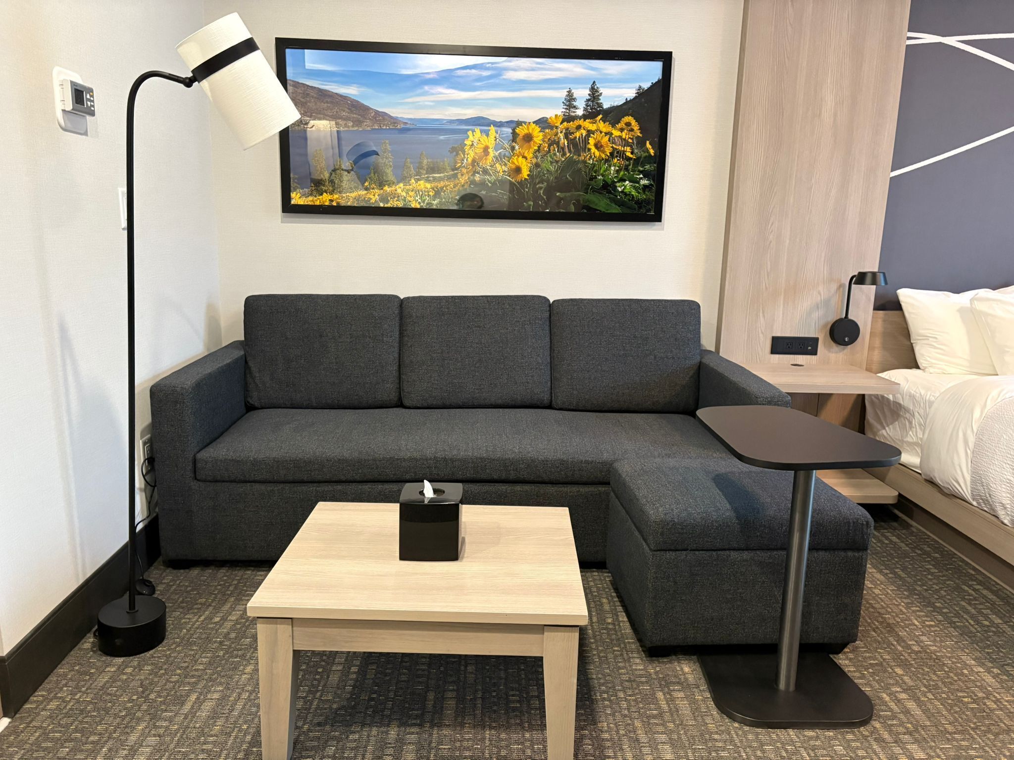 A modern hotel room with a gray sofa, a small wooden table, a bedside table, and a large TV mounted on the wall displaying a scenic landscape.