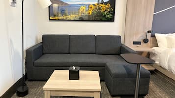 A modern hotel room with a gray sofa, a small wooden table, a bedside table, and a large TV mounted on the wall displaying a scenic landscape.