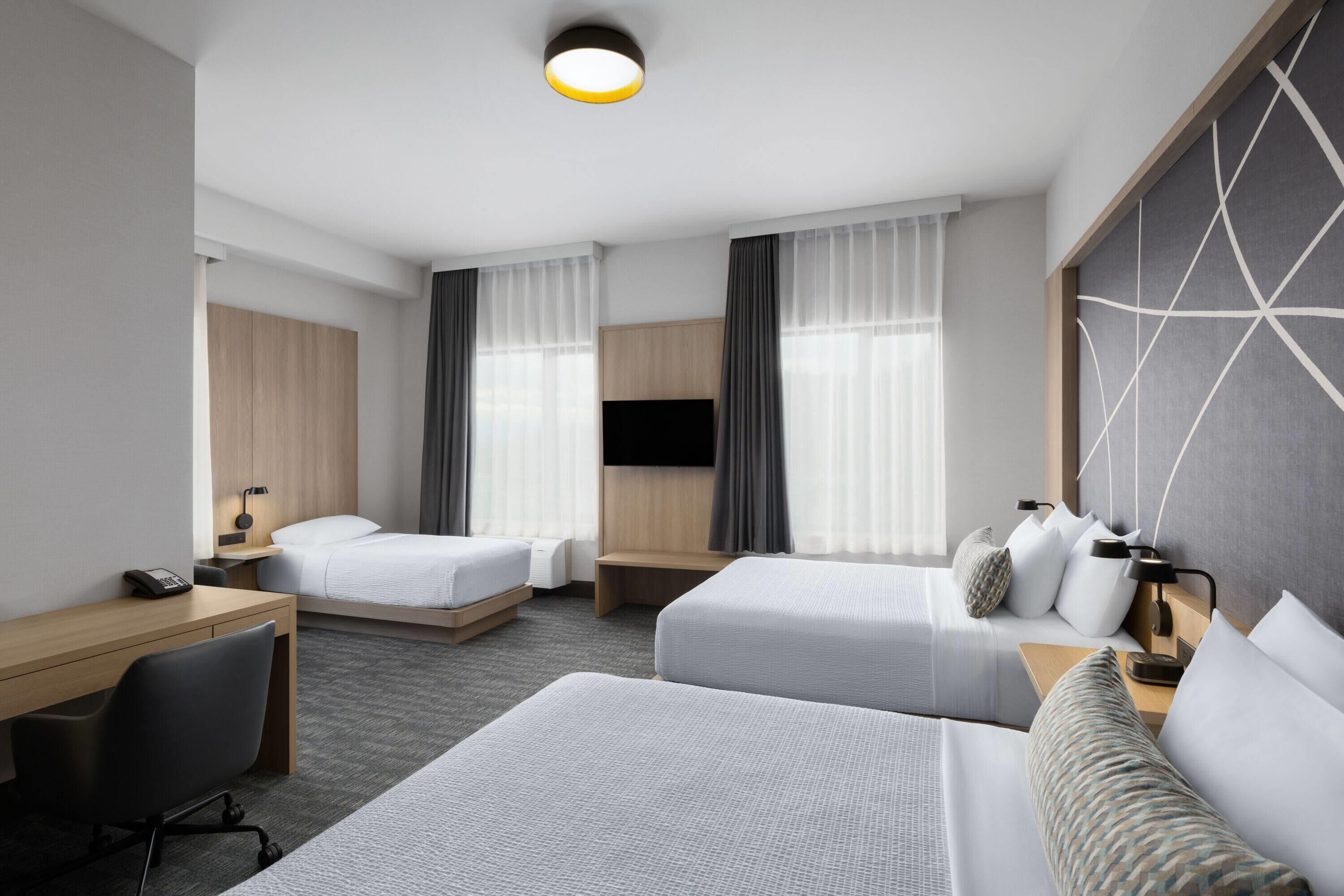 A hotel room with two beds, a desk, a chair, a TV, and a large window with curtains.