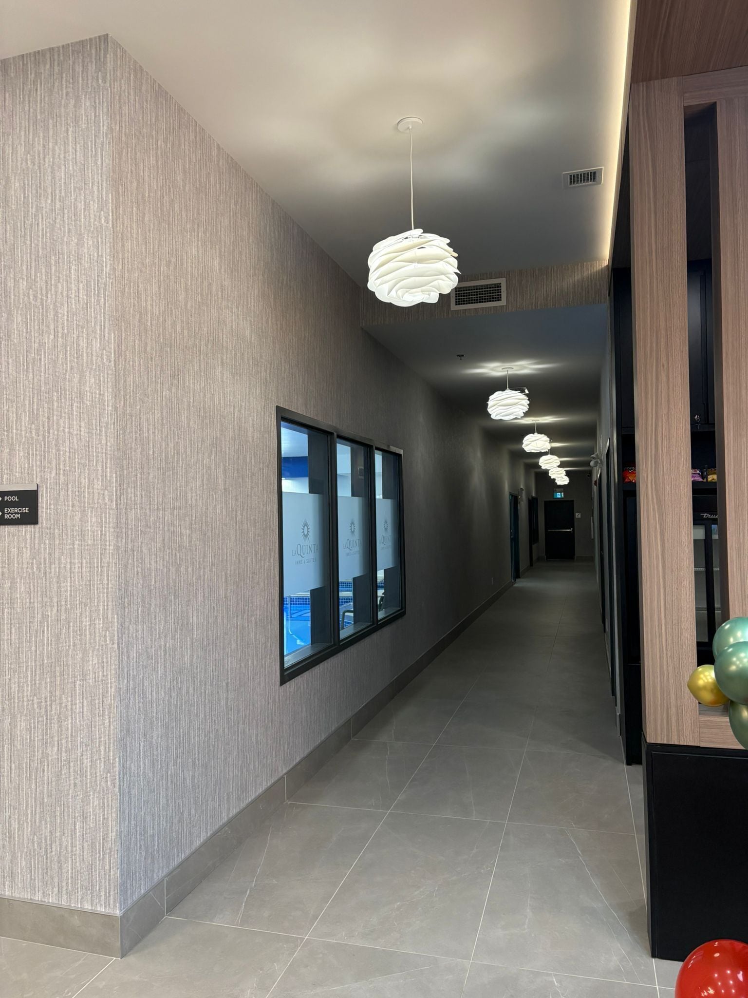 A modern hotel corridor with grey textured walls, a tiled floor, and multiple hanging pendant lights.