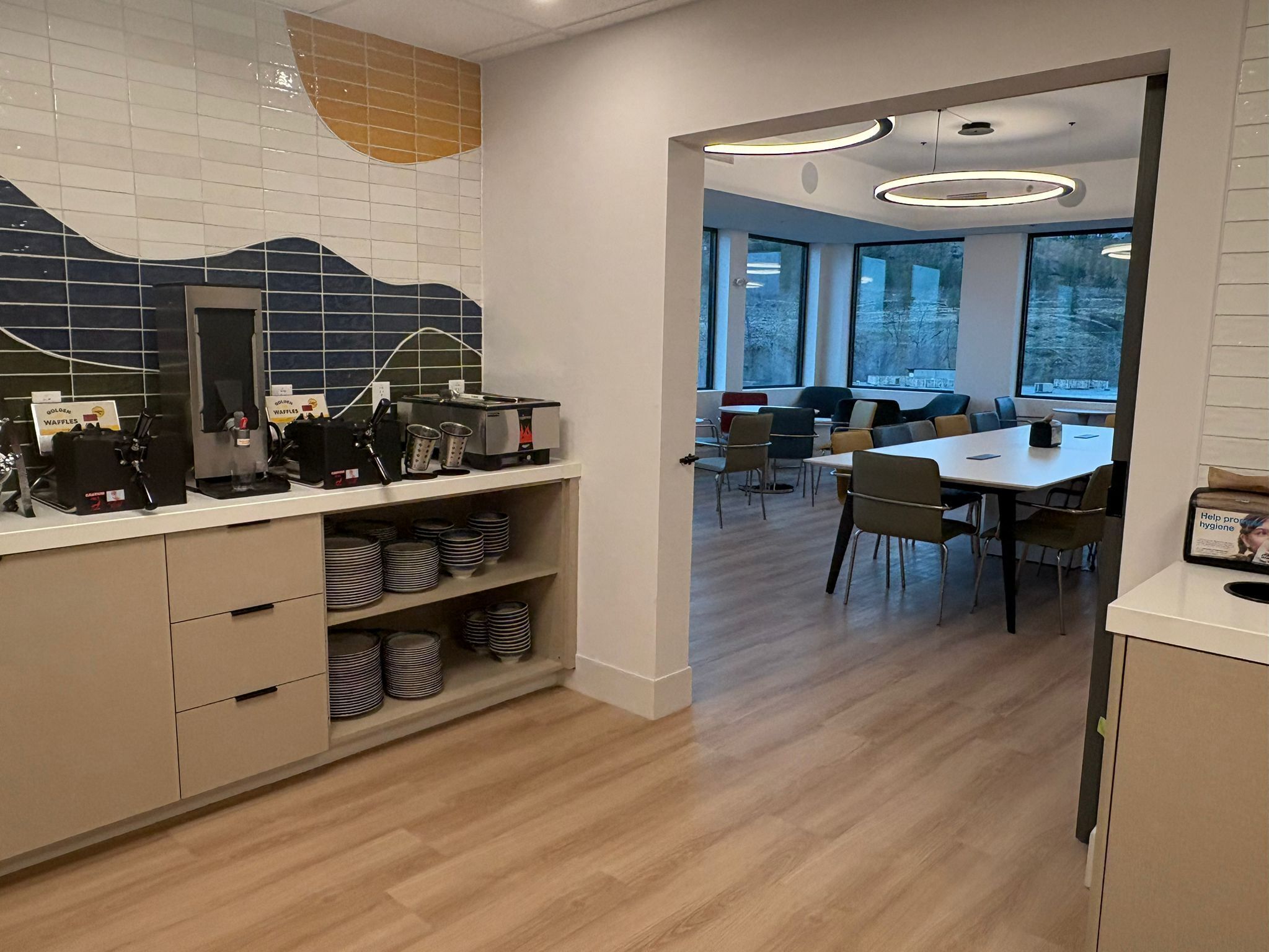 Uma cozinha moderna com estação de waffles, área de refeições com uma mesa grande e cadeiras, e vista para o exterior através de amplas janelas.