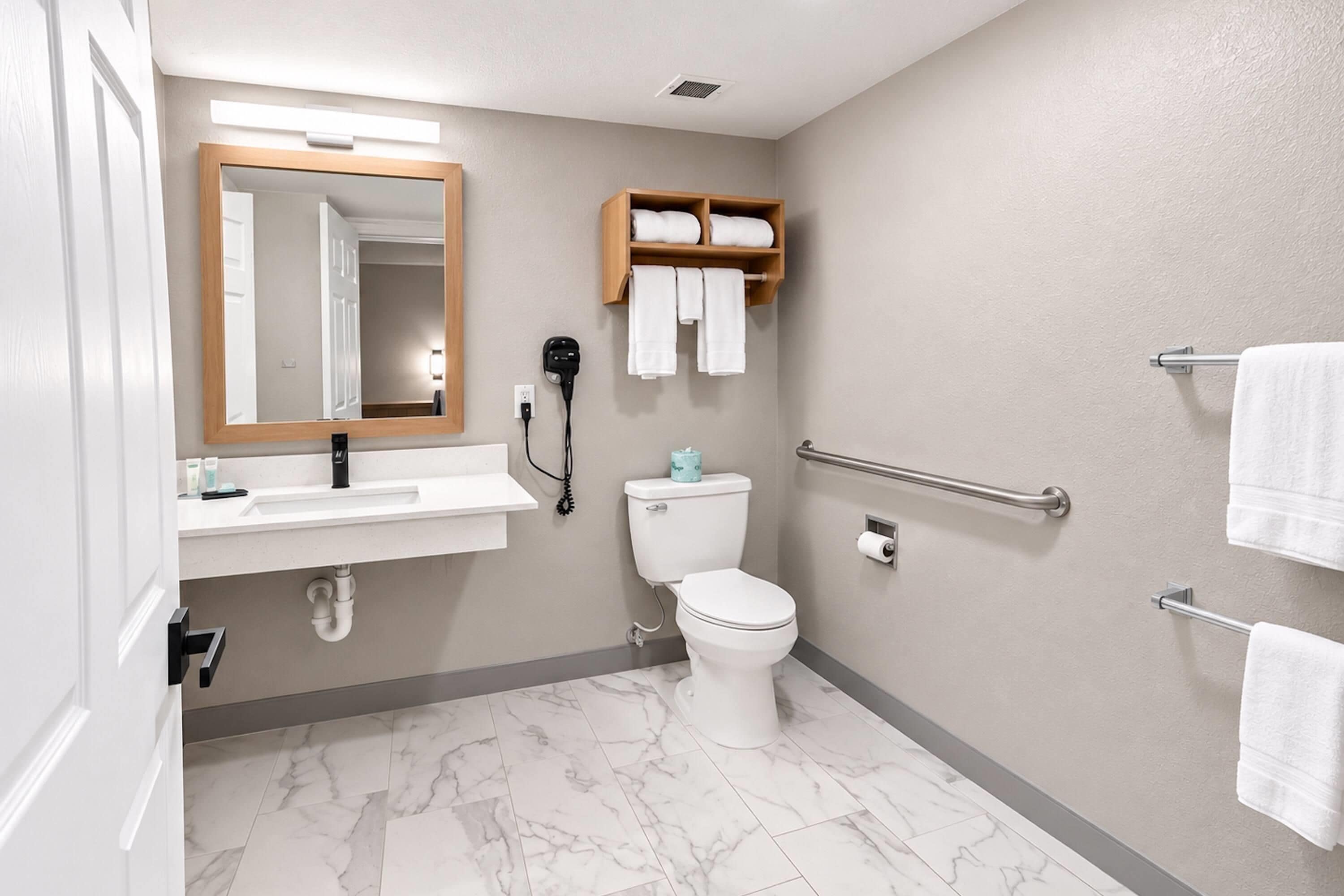 A bathroom with a sink, mirror, toilet, towel rack, and a handrail.