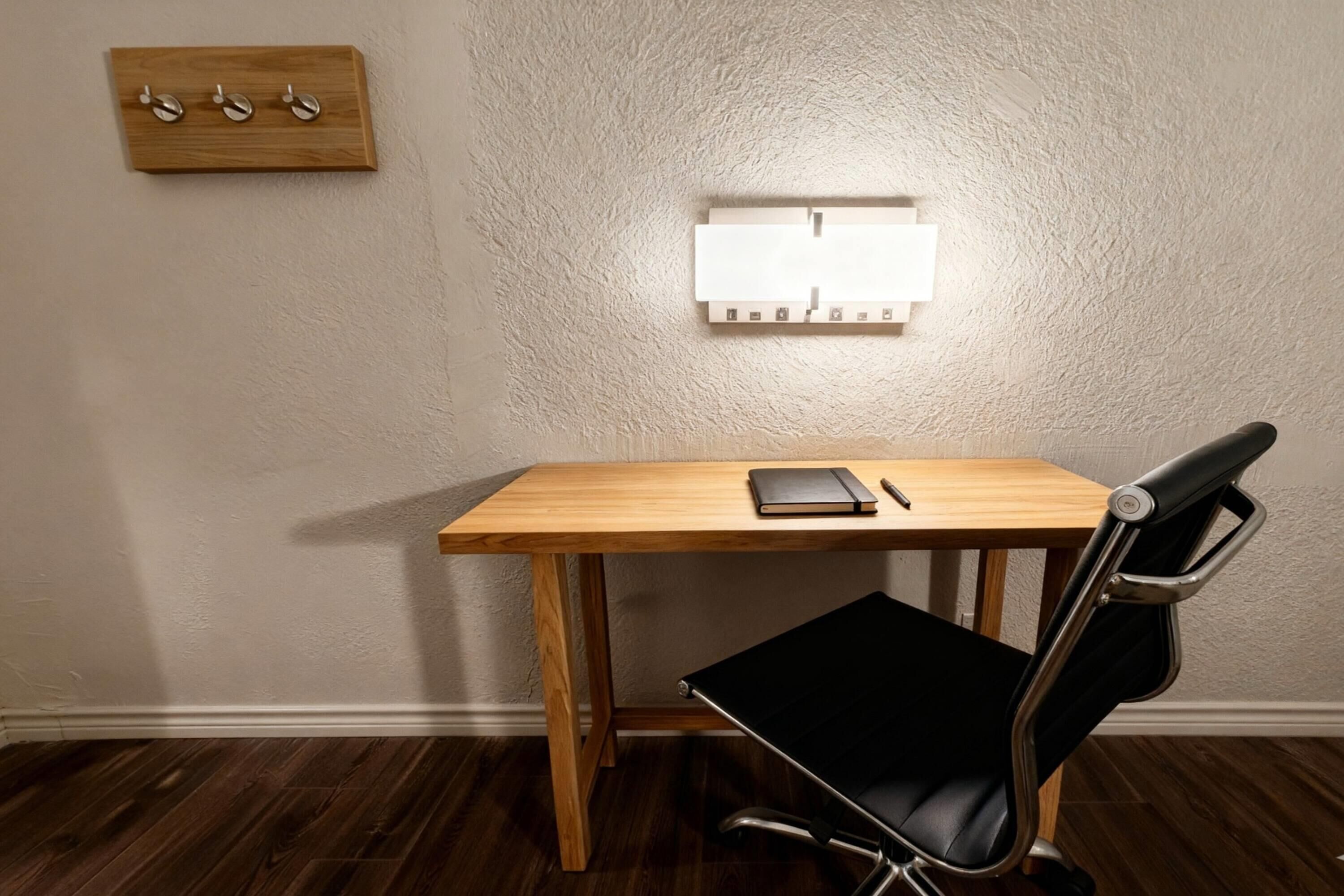A wooden desk with a black chair, a wall-mounted light switch, and a small wooden shelf with hooks.