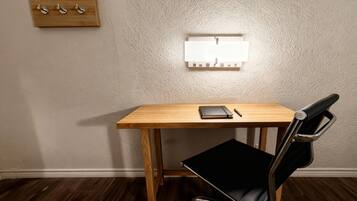 A wooden desk with a black chair, a wall-mounted light switch, and a small wooden shelf with hooks.