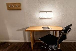 A wooden desk with a black chair, a wall-mounted light switch, and a small wooden shelf with hooks.