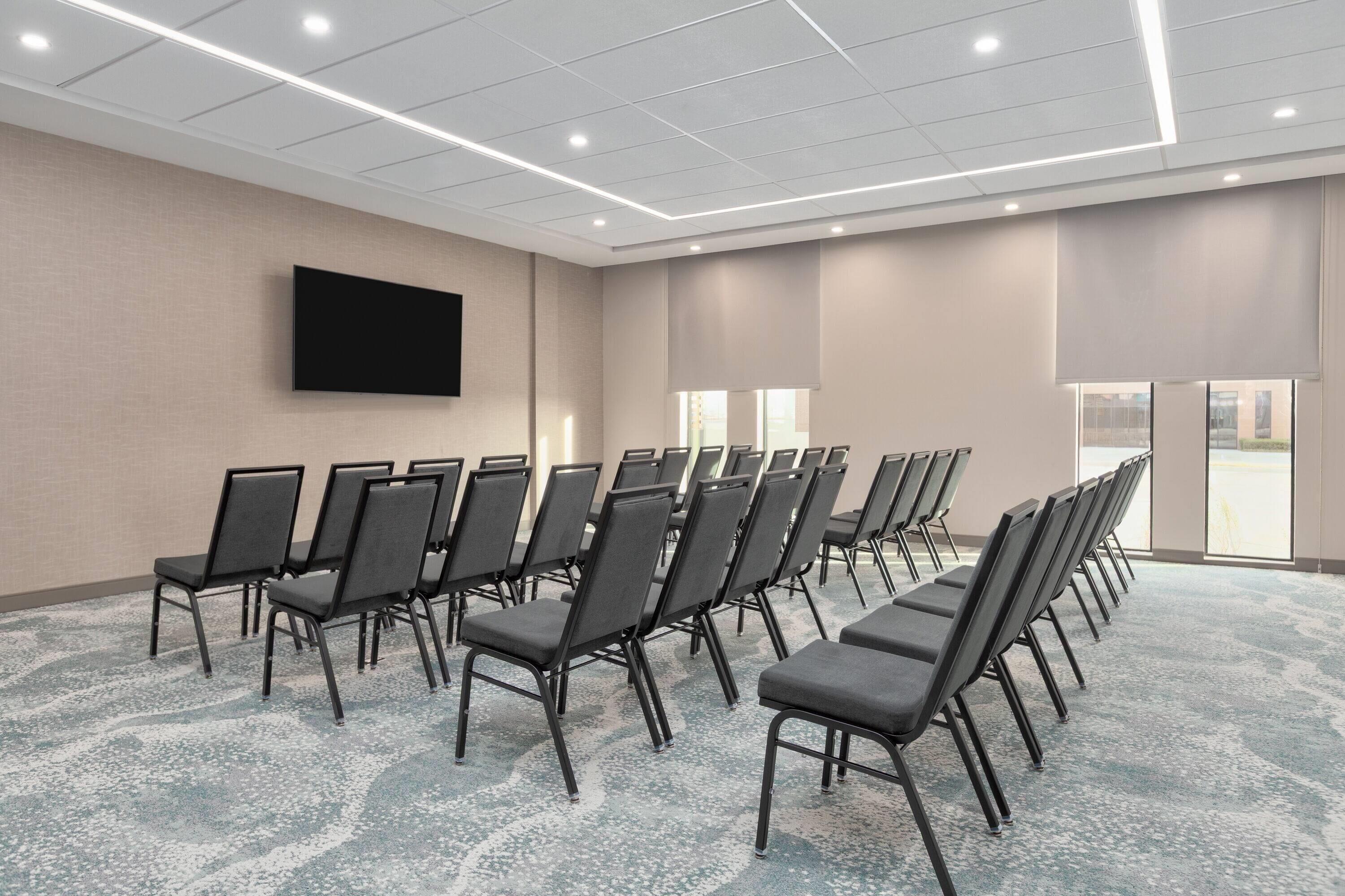 A conference room with rows of chairs, a flat-screen TV mounted on the wall, and large windows allowing natural light.