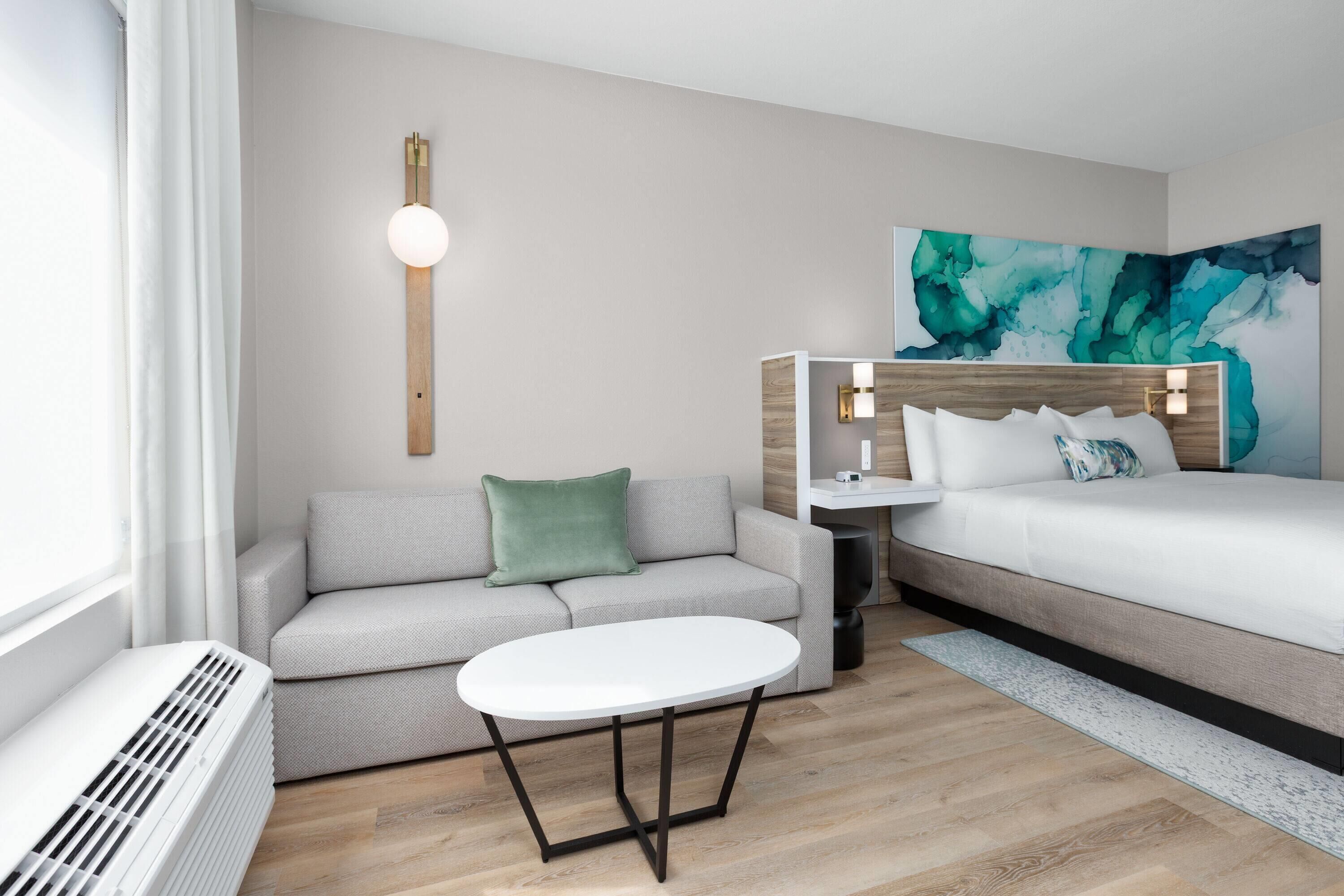 A modern hotel room with a grey sofa, a white table, a bed with a headboard, and a wall-mounted light fixture.