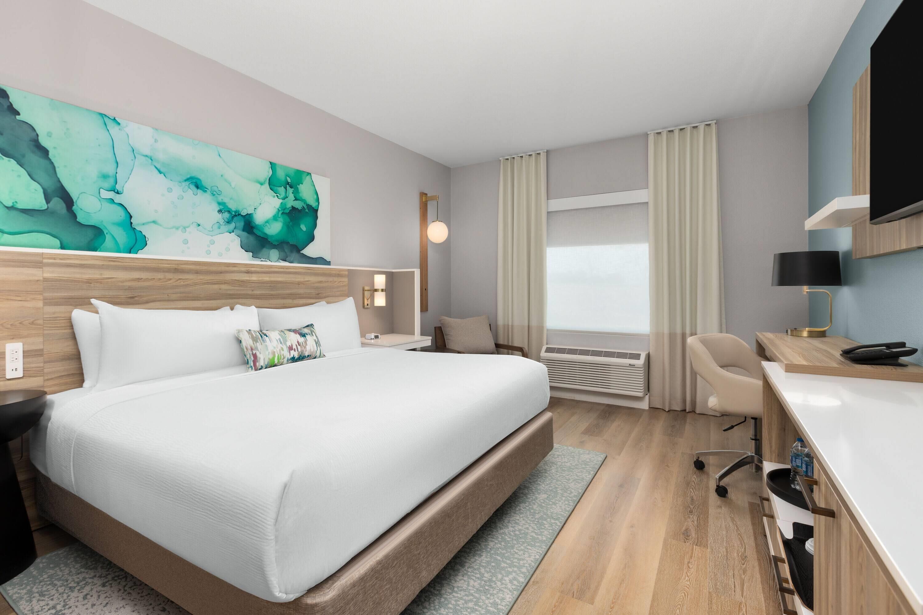 Habitación de hotel con una cama grande, un escritorio con silla, televisor y ventana con cortinas.