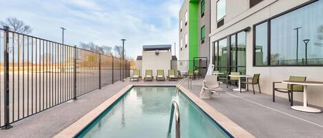 A swimming pool with a metal fence, outdoor seating, and a building with large windows.