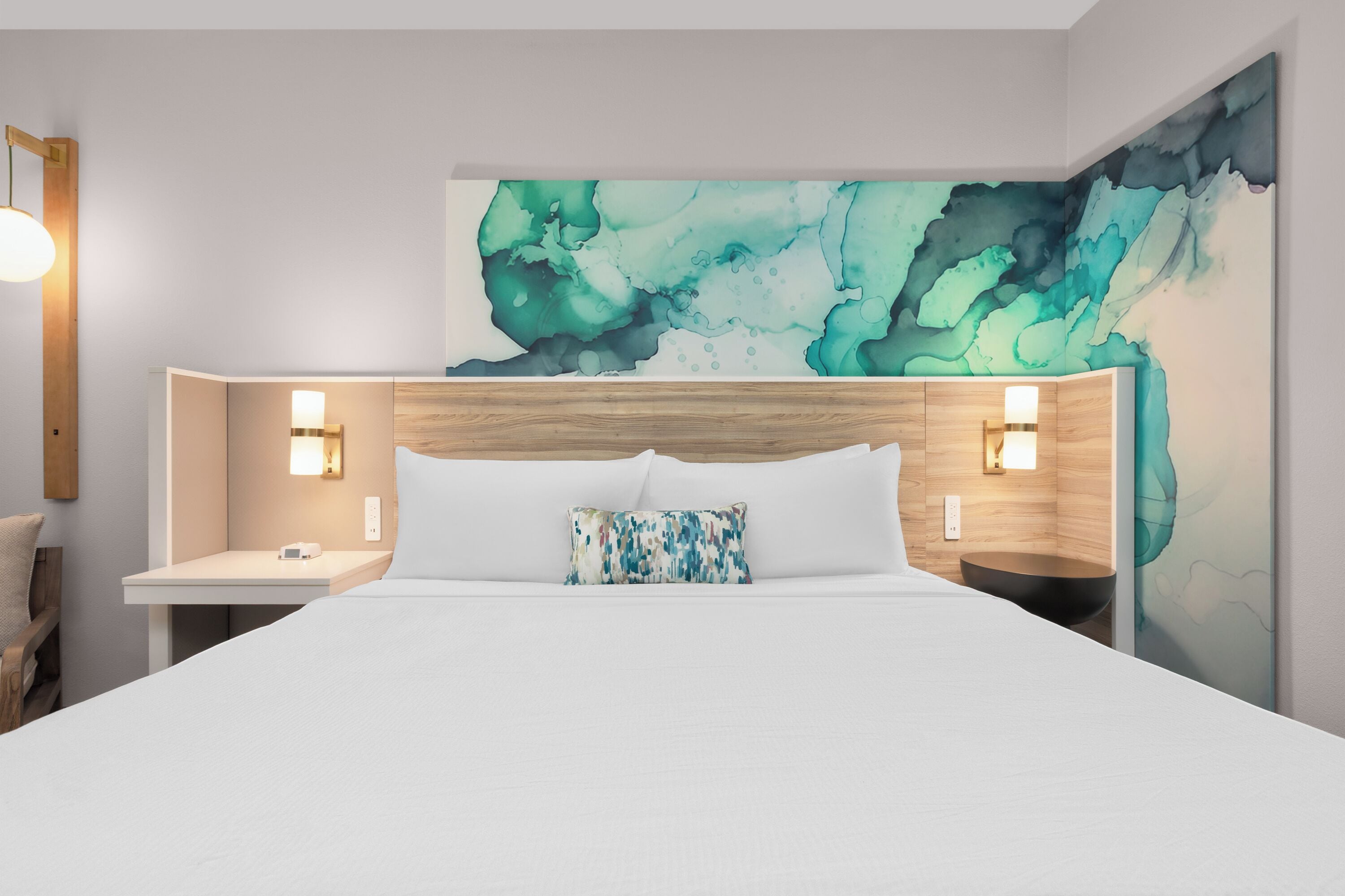 Una habitación de hotel moderna con una cama grande, dos mesitas de noche con lámparas, un cuadro abstracto de gran tamaño en la pared y un cabecero de madera.