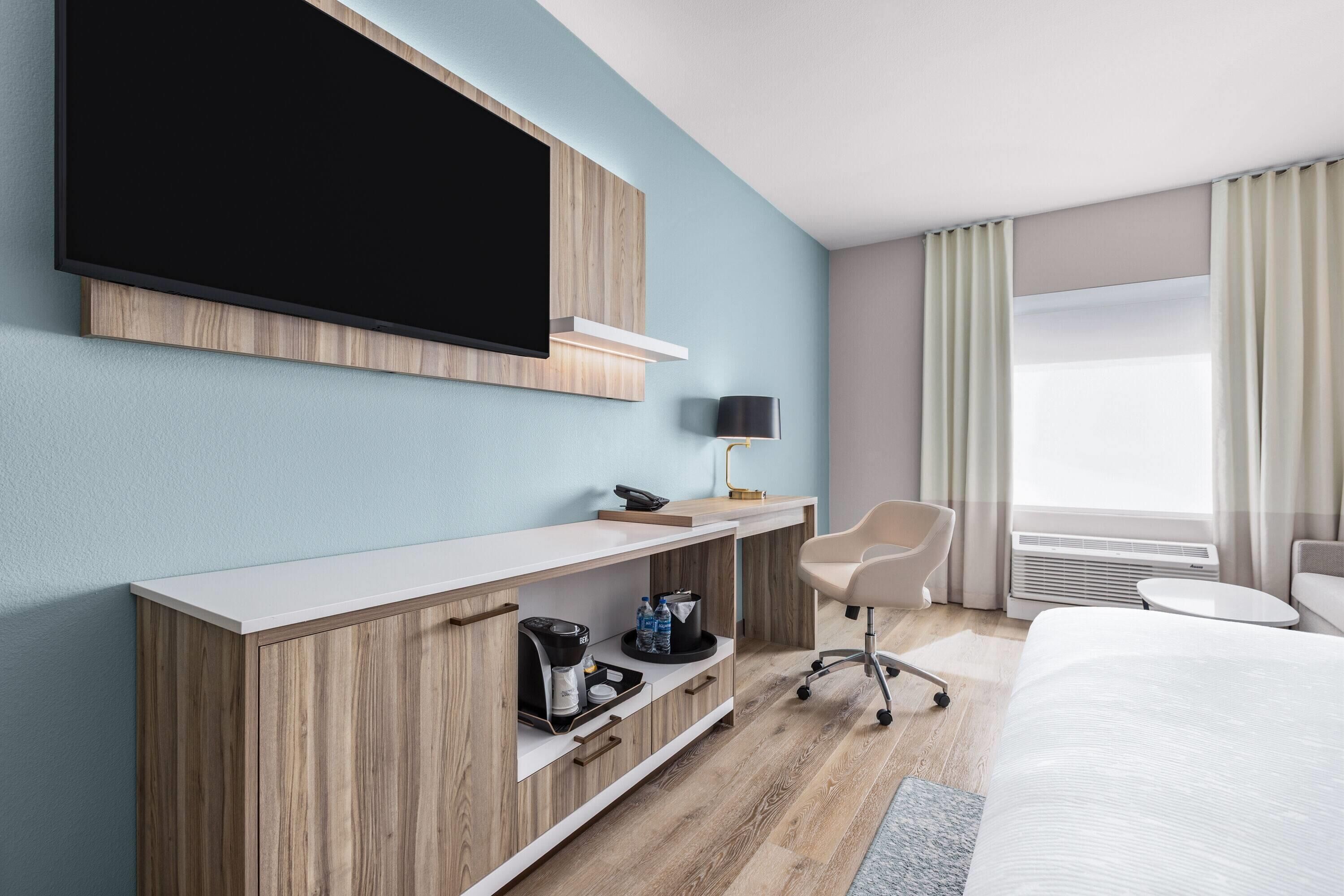 Habitación de hotel con cama, escritorio, televisor de pantalla plana, silla, cafetera y ventana con cortinas.