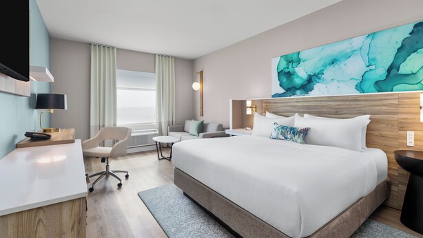 Ett hotellrum med en stor säng, ett skrivbord, en stol, en tv och en tavla på väggen.