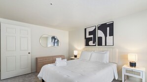Hotellihuone, jossa on sänky, yöpöytä, lamppu, peili ja seinätaideteos.