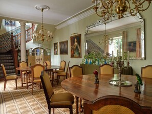 Comedor con una mesa de madera, sillas y una araña. Se ve una escalera con barandilla decorativa y un cuadro en la pared.