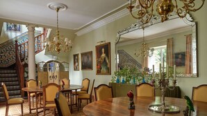 A dining room with a large wooden table, chairs, and a chandelier. There is a staircase with a decorative railing and a painting on the wall.
