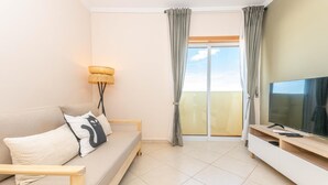 A modern living room with a sofa, a TV, a lamp, and a balcony with curtains. - Azura Apartment with 2 Bedrooms (Conceição de Tavira)