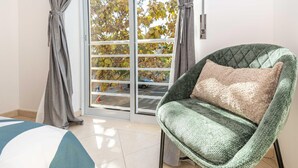 A room with a green armchair, a striped bedspread, and a sliding glass door leading to a balcony with autumn foliage. - Azura Apartment with 2 Bedrooms (Conceição de Tavira)