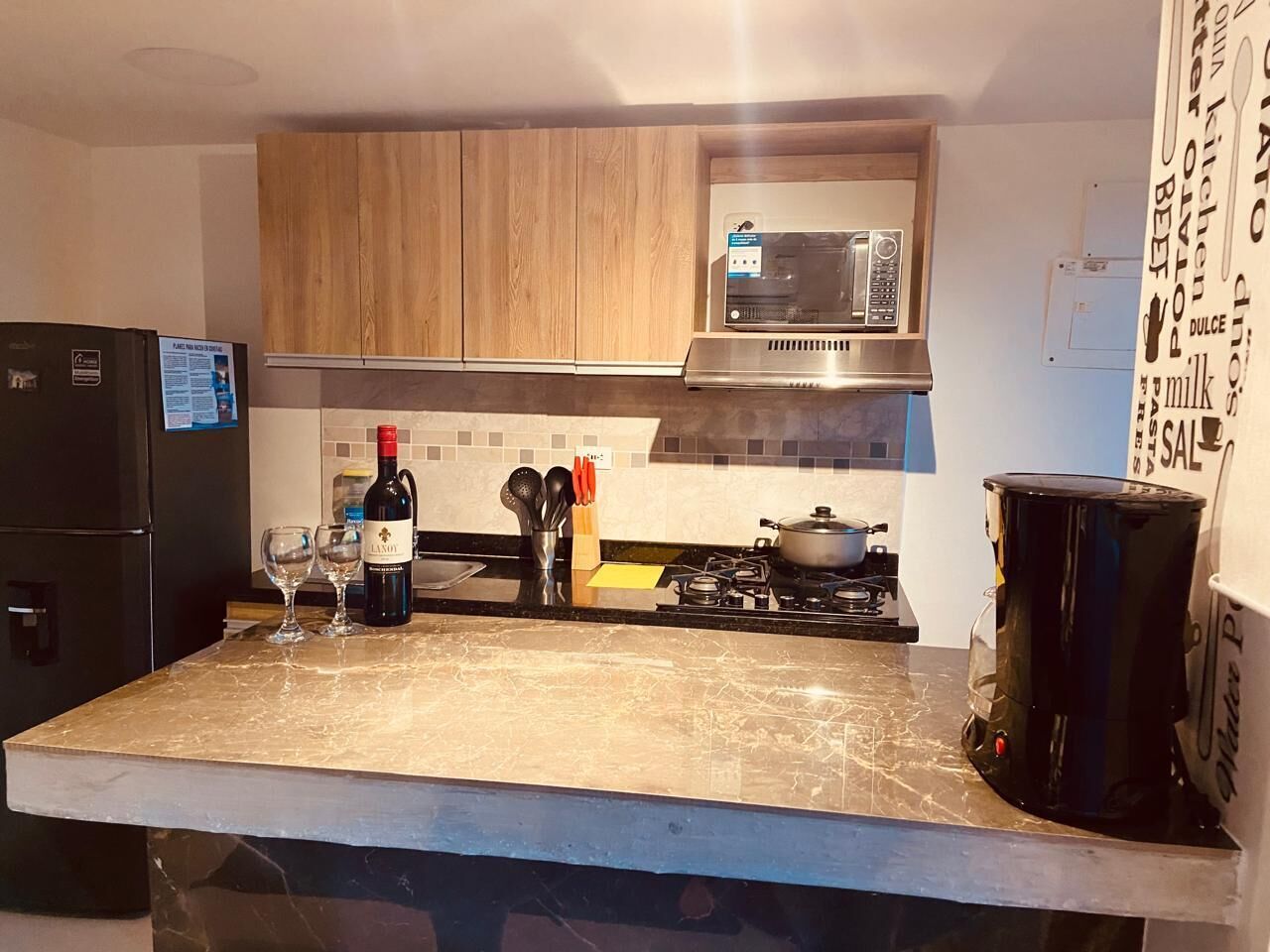 A kitchen with a refrigerator, wine bottle, glasses, a microwave, a stove, and a coffee maker.
