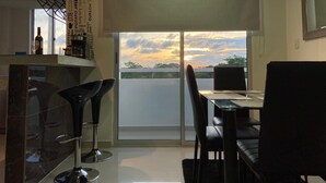 A modern kitchen with a dining area, a large window with a view of the sky, and a balcony.