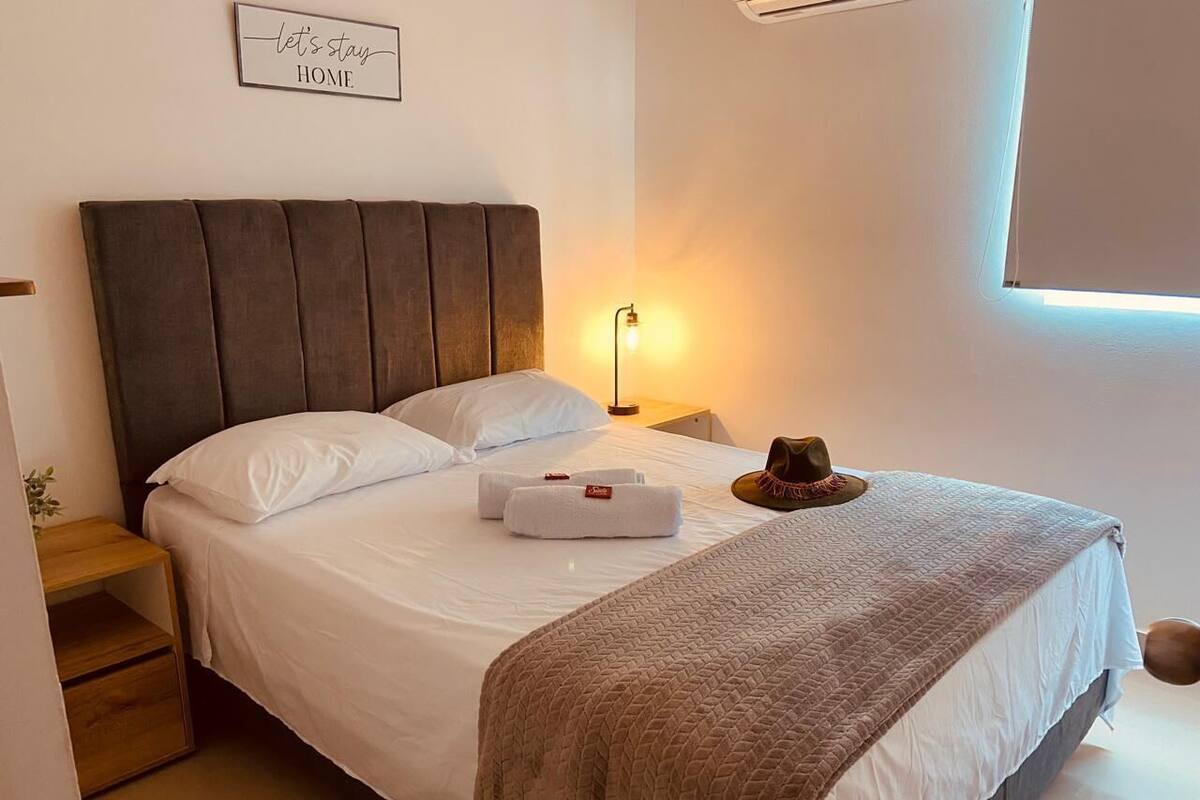 A neatly made bed with a headboard, a bedside lamp, and a hat on the bed.