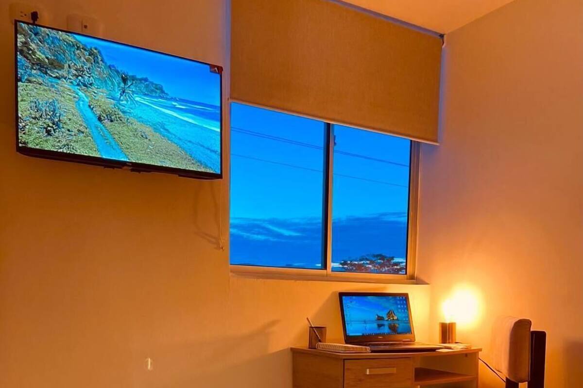 A hotel room with a bed, a desk with a laptop, a TV mounted on the wall, and a window with a view of the beach.
