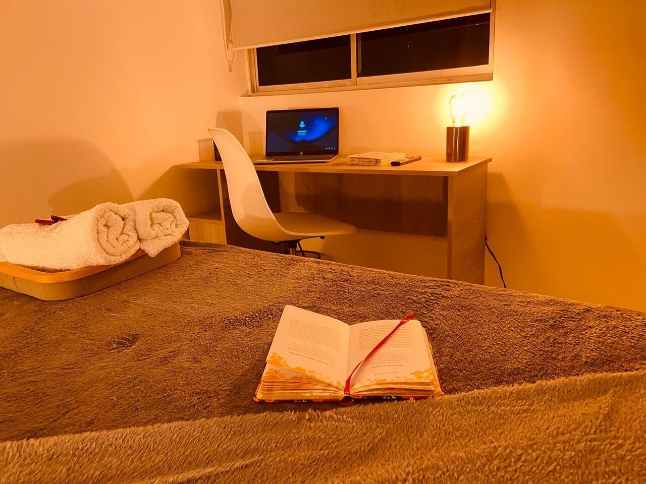 A room with a bed, a desk with a laptop, a chair, and a bedside table with a lamp.