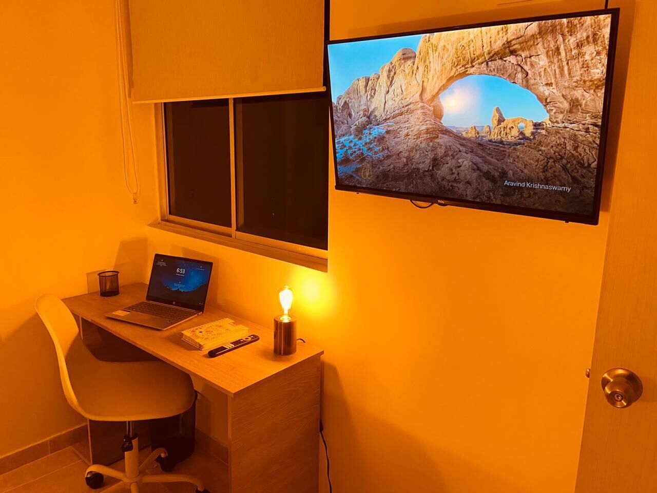 Una habitación con escritorio, computadora portátil, vela y televisor mostrando un paisaje.