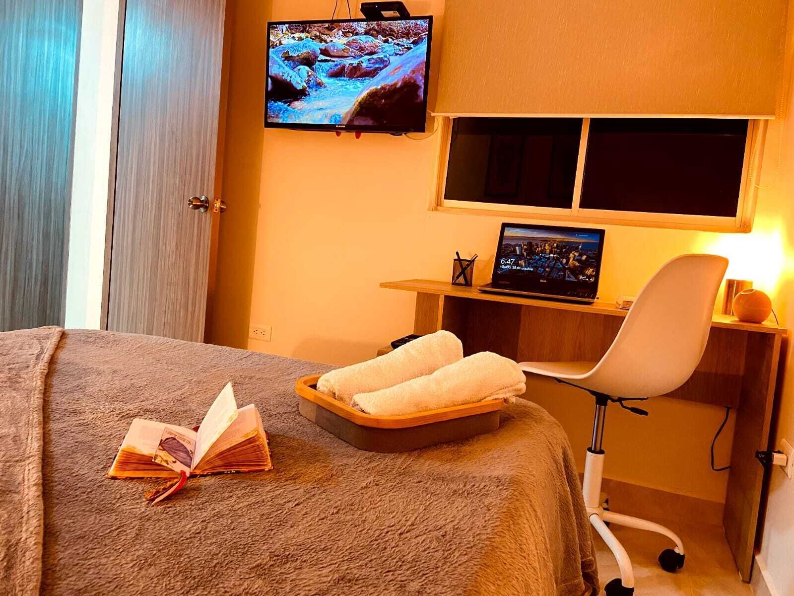 A hotel room with a bed, a desk with a laptop, a TV, and a chair.