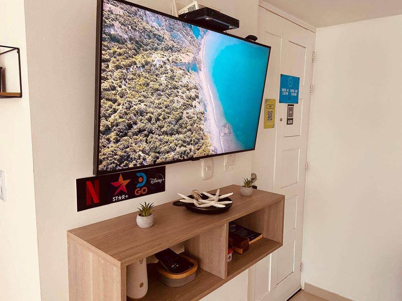 Una habitación de hotel moderna con un televisor de pantalla plana montado que muestra un paisaje pintoresco, una mesa consola de madera con adornos, y una puerta con un letrero azul que indica disponibilidad.