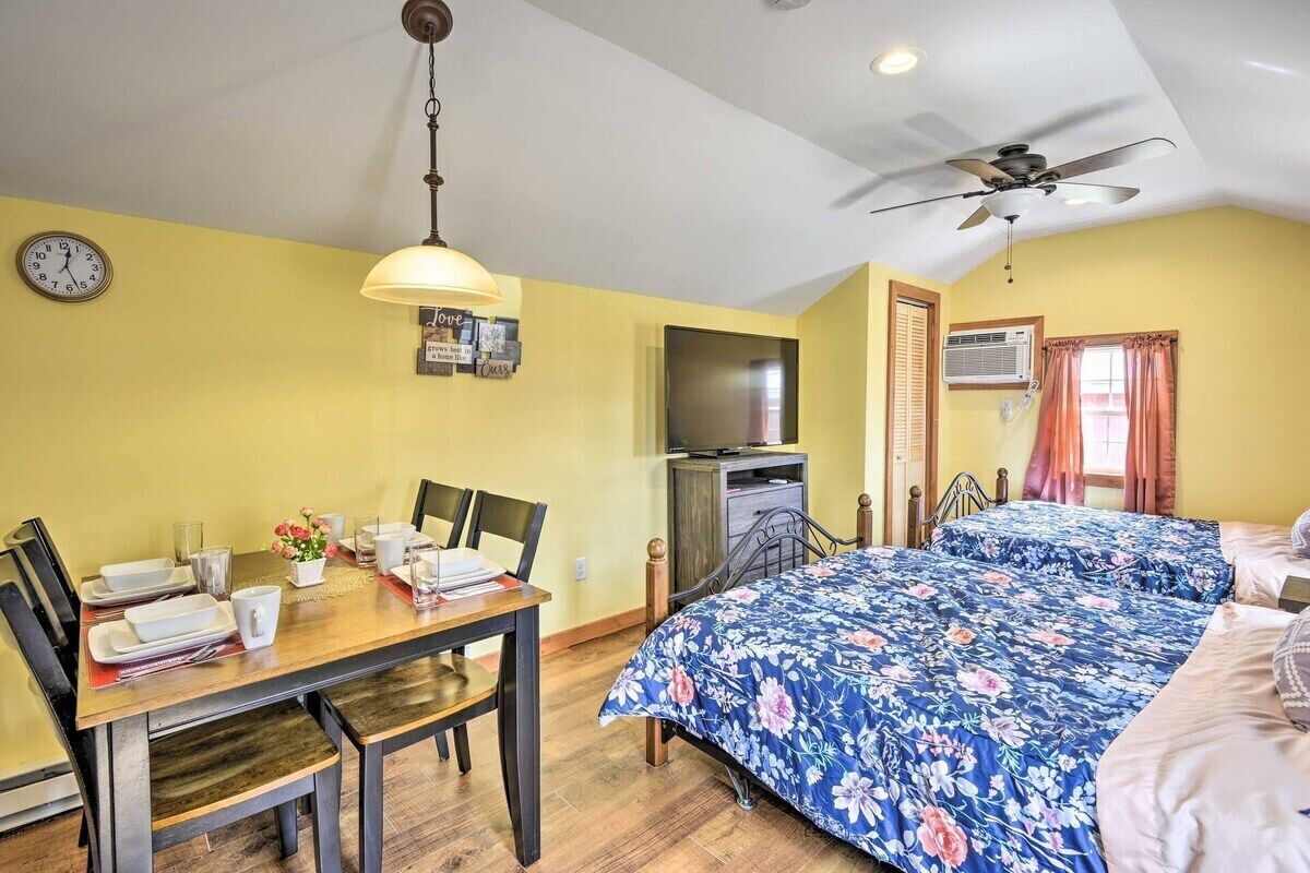 A room with a bed, a TV, a dining table with chairs, a clock, and a ceiling fan.