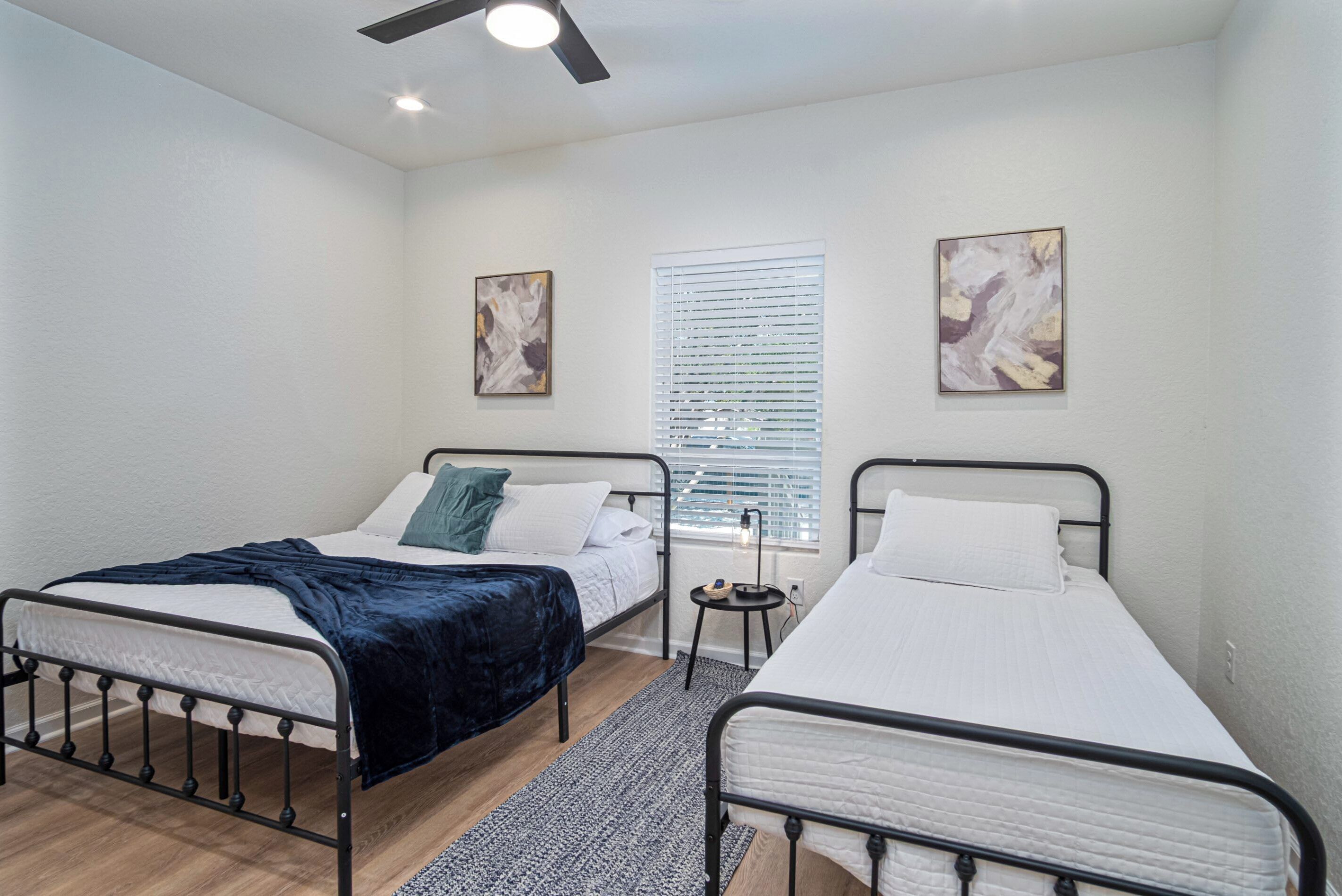 A bedroom with two single beds, a ceiling fan, a window with blinds, and two wall-mounted paintings.