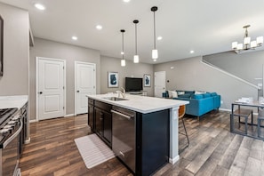 Una cocina moderna con una isla central, un comedor con mesa y sillas, una sala de estar con sofá, y una escalera que conduce a un nivel superior.