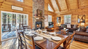 L’intérieur chaleureux d’un chalet en rondins, avec une cheminée en pierre, des meubles en bois et de grandes fenêtres offrant une vue sur la forêt environnante.