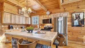 Une cuisine rustique dans une cabane en bois, avec un îlot central en pierre, des armoires en bois, un lustre, un four à micro-ondes, un réfrigérateur et un tableau représentant un ours.